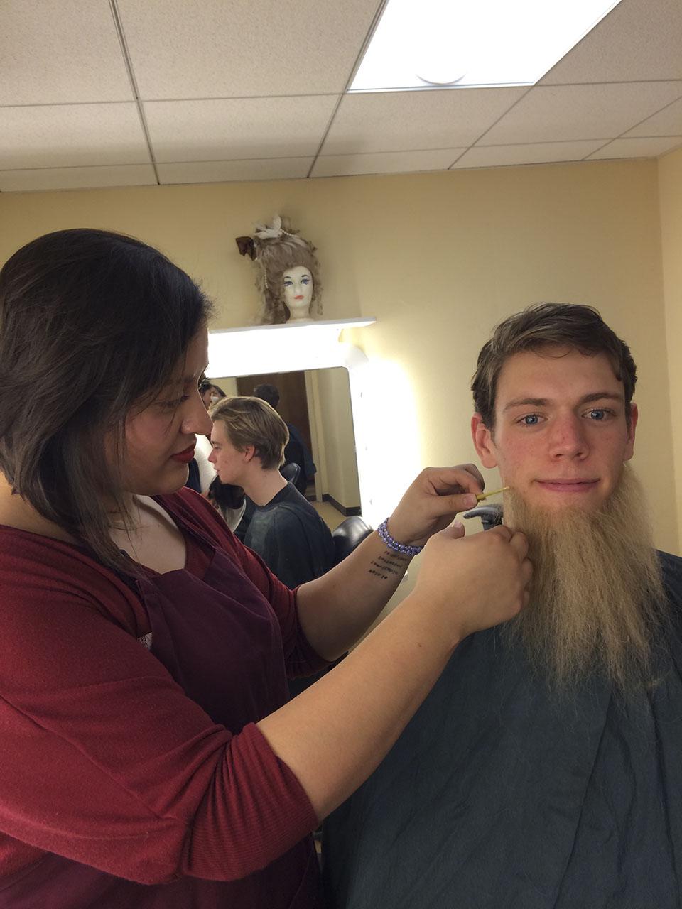A female student with dark hair applies a false beard to a caucasian male student.