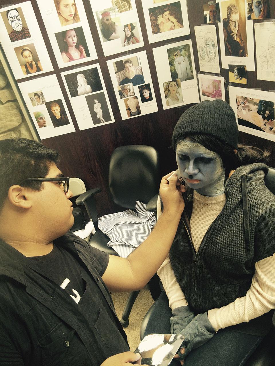 A male student with dark hair applies a monster gray character makeup to a student wearing a black beanie. Makeup images can be seen on the wall behind them.