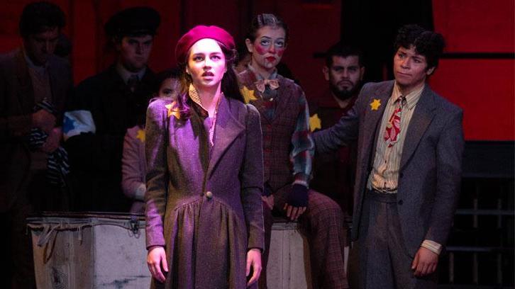 An actress in a red beret and coat is illuminated center stage as actors in suits look on.